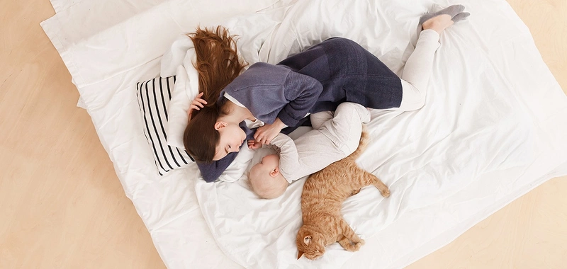 En mor ligger på gulvet på pledd med baby og en katt.