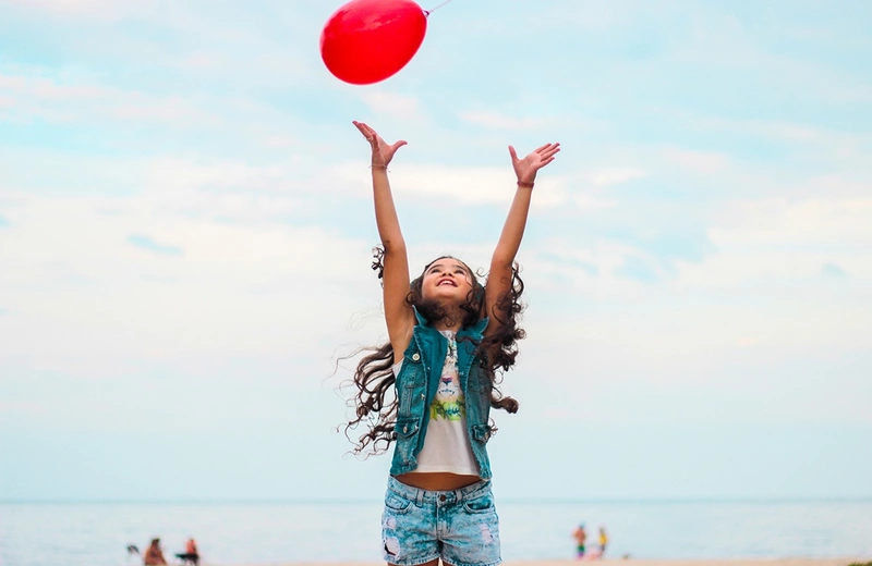 Jente strekker seg etter rød ballong på en strand. Foto