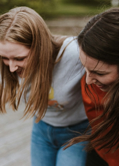Bilde av to smilende, unge kvinner som holder en arm rundt skuldrene på hverandre. Håret blafrer som om de er i bevegelse. 